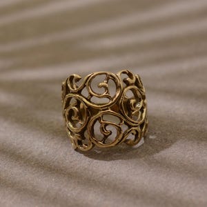 May include: A gold-coloured ring with an openwork design featuring swirling, ornate patterns. The ring is a wide band style, with the decorative elements covering the entire surface. The ring is set against a neutral background.