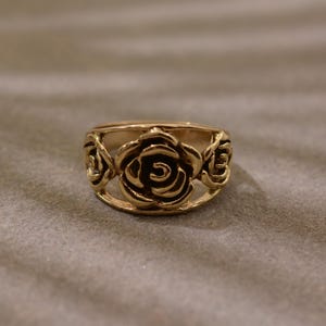 May include: A gold-colored ring featuring a floral design. The ring has three openwork rose motifs evenly spaced around the band. The roses are detailed with visible petals and a central spiral.