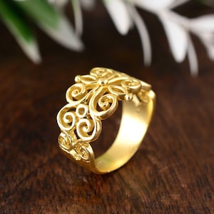 Può includere: Un anello color oro con un intricato design traforato. L'anello presenta un motivo floreale e a volute, con una finitura lucida. L'anello è mostrato su una superficie di legno scuro.