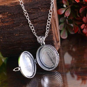 May include: Silver locket necklace with an oval pendant, open to show the interior. The pendant is attached to a delicate chain. The locket is open, showing the inside. The background is a dark wooden surface.