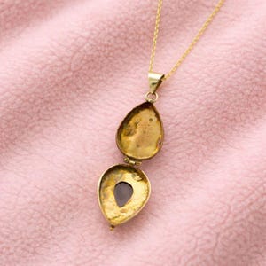 Peut inclure: Un collier avec un pendentif en forme de goutte d'eau doré, ouvert pour révéler un intérieur sombre. Le pendentif est suspendu à une fine chaîne dorée. Le médaillon est posé sur un fond rose tendre.