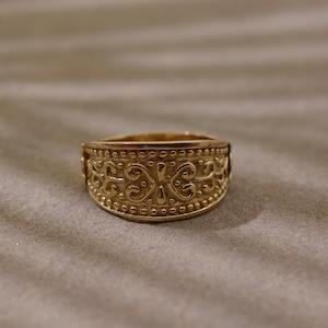 May include: A gold-coloured ring with an ornate design. The ring features a band with a repeating pattern of swirls, dots, and floral-like shapes. The ring is likely made of metal and has a polished finish. The ring is a piece of jewellery.