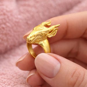May include: A gold ring with a goat head design. The ring is held in a hand against a pink background. The goat's head has textured details, including horns and facial features. The ring is a piece of jewellery with a unique animal theme.