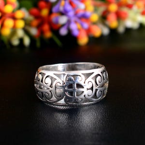 Puede incluir: Un anillo plateado con un diseño calado con patrones arremolinados y en forma de cruz. El anillo está sobre un fondo oscuro con flores coloridas borrosas en el fondo.