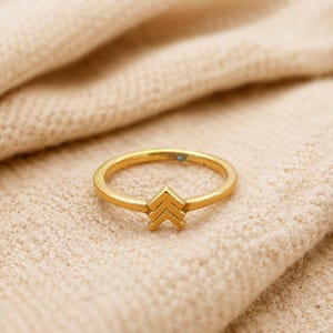 Peut inclure: Une bague dorée avec un motif de chevrons. La bague est posée sur un tissu de couleur neutre. La bande est fine, et le chevron est composé de trois chevrons empilés pointant vers le haut.