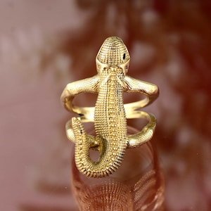 Pode incluir: Um anel dourado em forma de lagarto. O lagarto tem um corpo texturizado e envolve o dedo. O anel é feito de um material metálico brilhante e tem um design detalhado.