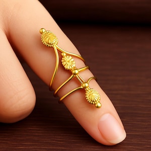 Puede incluir: Un anillo de dedo dorado con un diseño floral. El anillo tiene tres bandas que envuelven el dedo, conectadas por una barra vertical con elementos florales decorativos. El anillo está hecho de un material metálico brillante.