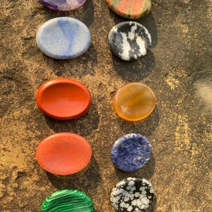 May include: An assortment of polished, oval-shaped gemstones in various colors, including purple, green, red, blue, and black. The stones are arranged on a textured, brown surface. The gemstones have unique patterns and textures.