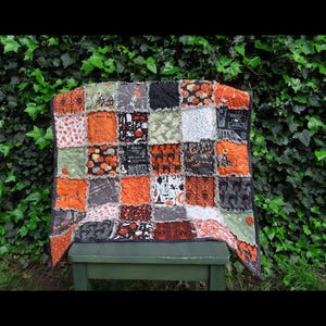 Puede incluir: Un edredón de Halloween de patchwork con cuadrados naranjas, negros, grises y verdes. El edredón presenta varios patrones temáticos de Halloween, incluyendo calabazas, murciélagos y fantasmas. El edredón se exhibe sobre un banco verde.