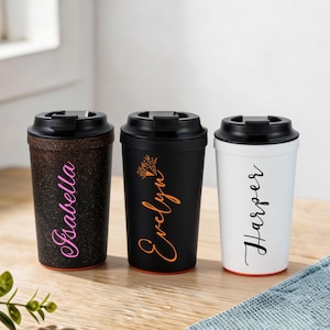 Puede incluir: Tres tazas de viaje reutilizables con tapas negras. La taza de la izquierda es de color marrón oscuro con textura moteada y el nombre Isabella en rosa. La taza central es de color negro mate con un gráfico de fuegos artificiales y el nombre Evelyn en naranja. La taza de la derecha es blanca con el nombre Harper en negro.