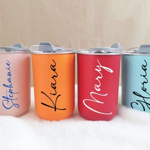 Puede incluir: Cuatro vasos térmicos de colores con nombres: Stephanie, Kiara, Mary y Gloria. Los vasos son en tonos rosa, naranja, rojo y azul, cada uno con una tapa a juego. Los nombres están escritos en cursiva.