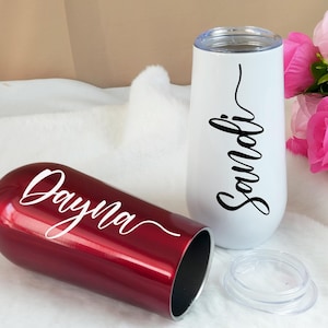 Op de afbeelding: Twee geïsoleerde champagnefluiten zonder steel. Eén is glanzend rood met de naam "Dayna" in witte letters. De andere is wit met de naam "Sandi" in zwarte letters. Een doorzichtig deksel ligt in de buurt. De achtergrond bevat roze bloemen.