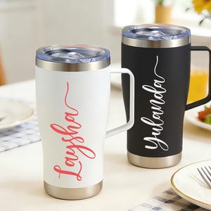 May include: Two insulated travel mugs with handles. One is white with "Laysha" in red script, the other is black with "Yulanda" in white script. Both have silver bases and clear lids. They sit on a table with a plate and cutlery.