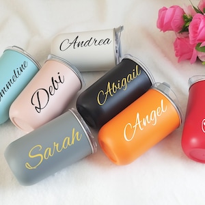 Puede incluir: Una colección de vasos personalizados en varios colores, incluyendo azul, rosa, gris, negro, naranja, blanco y rojo. Cada vaso tiene un nombre impreso en un color contrastante. Los vasos están dispuestos sobre una superficie blanca.