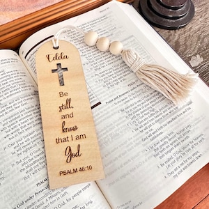 May include: A wooden bookmark with the text "Be still, and know that I am God" and a cross cutout. It has a tassel and wooden beads attached. The bookmark is resting on an open book.