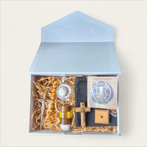 Puede incluir: Una caja de regalo azul claro que contiene artículos religiosos. En el interior hay un rosario, un crucifijo, una medalla de San Benito, una cruz de madera, una botella pequeña y un folleto. La caja está llena de papel triturado.