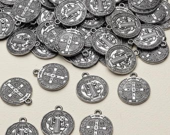 May include: A pile of silver Saint Benedict medals. Each circular pendant displays religious iconography and text, including a cross and the letters "NDSMD". The medals have a loop for attaching to a chain or cord.
