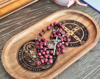 Puede incluir: Un rosario de madera con cuentas granates y un crucifijo plateado descansa sobre una bandeja ovalada de madera. La bandeja presenta símbolos y texto religiosos grabados. El rosario es un artículo devocional católico tradicional.