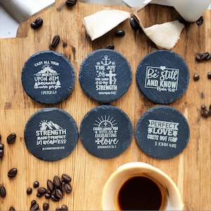 Puede incluir: Seis posavasos redondos de pizarra gris oscuro con citas inspiradoras y versículos bíblicos grabados en blanco. Los posavasos están dispuestos sobre una superficie de madera con granos de café y una taza de café.