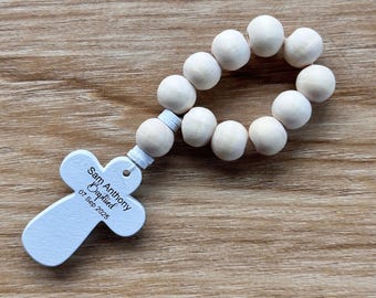 Puede incluir: Un rosario de madera con cuentas grandes y redondas de color claro. El colgante en forma de cruz es blanco y tiene la inscripción "Sam Anthony Baptized 07 Sep 2025". El rosario se muestra sobre una superficie de madera.