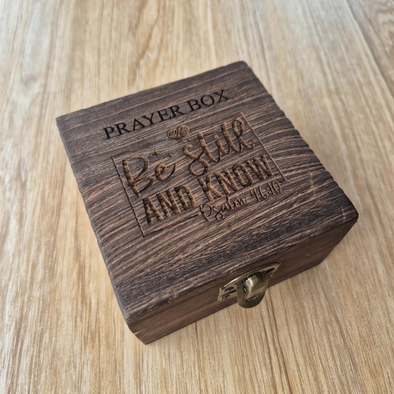 Puede incluir: Caja de oraci&oacute;n de madera marr&oacute;n oscuro con tapa con bisagras y cierre met&aacute;lico. La caja est&aacute; grabada con las palabras "PRAYER BOX" y "Be Still AND KNOW" y un vers&iacute;culo b&iacute;blico. La madera tiene una veta texturizada.