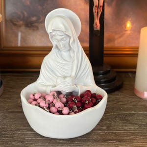 May include: A white ceramic statue of the Virgin Mary holding a heart-shaped bowl filled with pink and red rosary beads. The statue has a serene expression and is set against a blurred background.
