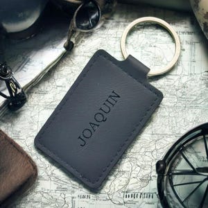 以下が含まれることがあります： 「JOAQUIN」の名前がエンボス加工された濃いグレーの革製キーホルダー。 キーホルダーは銀色のリングが付いており、ヴィンテージマップの背景に配置されています。 その他の旅行用アクセサリーも表示されています。