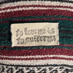 May include: A rectangular off-white fabric label with frayed edges featuring the black text "to love me is to suffer me" in a stylized font, stitched onto a woven textile background with horizontal stripes in maroon, dark green, and white.