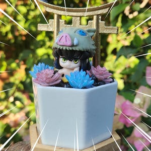 May include: A white square ceramic planter with a miniature anime figure wearing a boar head, surrounded by blue and pink artificial succulents. The planter sits on a wooden base, with a gold decorative arch in the background.