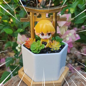 May include: A white hexagonal planter with a miniature anime figure, a golden torii gate, and small succulents. The planter sits on a wooden base. The figure has yellow hair and is holding a heart.
