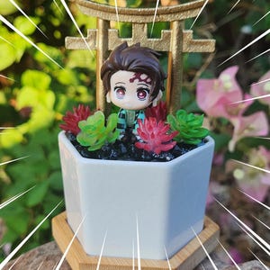 May include: A white hexagonal planter with a miniature anime figure, surrounded by red and green succulents. The figure is positioned in front of a gold-colored torii gate. The planter sits on a wooden base.