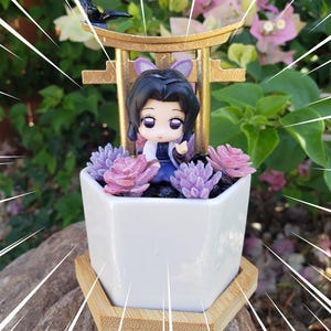 May include: A white ceramic planter with a miniature anime figure, surrounded by purple and pink faux succulents. The planter sits on a bamboo tray, with a gold-colored arch and a small black bird figurine.