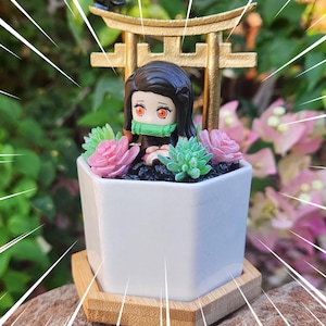 May include: A white ceramic planter featuring a miniature anime figure, pink and green faux succulents, and a golden torii gate. The planter is set on a wooden base, with the "Khaida Kreations" logo visible.
