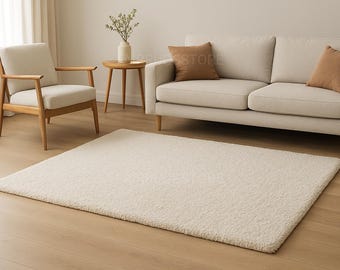 May include: A cream-colored, rectangular rug with a textured surface is the focal point of a living room scene. A light-colored sofa with two brown throw pillows, a wooden chair, and a side table with a vase of flowers complete the room.