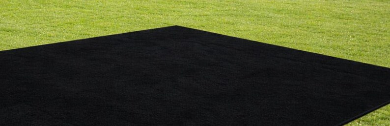 May include: A large, black rectangular rug is placed on a green grassy surface. The rug's dark color contrasts with the bright green grass, creating a simple yet striking visual. The rug appears to be made of a textured material.