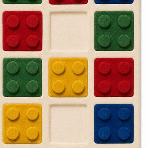 May include: A rectangular beige rug featuring a grid of square cutouts. Some squares contain raised, textured block shapes in red, blue, green, and yellow, each with four circular studs. Other squares are empty, showing the flat beige base.