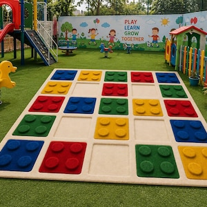 May include: A large rectangular floor mat featuring a grid of square indentations, some filled with raised, colorful, four-stud building block shapes in red, blue, green, and yellow. The mat is positioned on artificial grass in a playground.