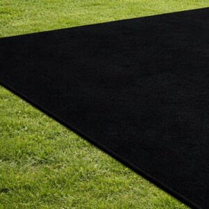 May include: A large, rectangular black rug is placed on a bed of green grass. The rug's dark color contrasts with the bright green of the grass, creating a striking visual effect. The rug appears to be made of a soft, textured material.