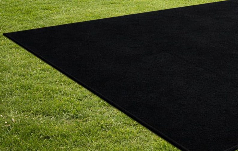 May include: A large, rectangular black rug is placed on a bed of green grass. The rug's dark color contrasts with the bright green of the grass, creating a striking visual effect. The rug appears to be made of a soft, textured material.