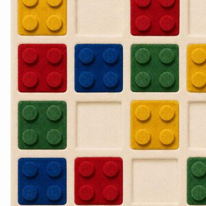 May include: A square rug featuring a grid pattern with raised, textured blocks in primary colors including red, blue, yellow, and green. Each block has four circular studs on its surface, set against a neutral, cream-colored background.