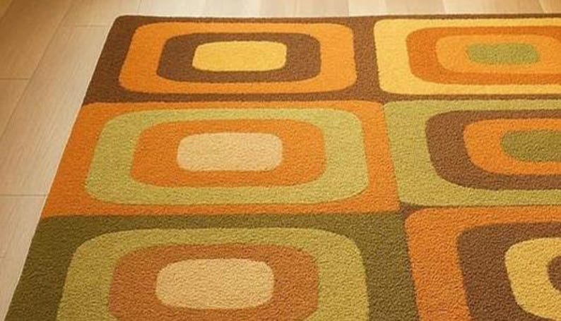 This rug features a retro geometric design with bold, rounded square patterns in warm shades of orange, mustard yellow, olive green, and brown. Its soft texture and mid-century modern style make it a standout piece for any living room or bedroom. The combination of earthy tones and clean, vintage-inspired shapes gives the rug a cozy yet stylish appeal — perfect for adding warmth and personality to your space.