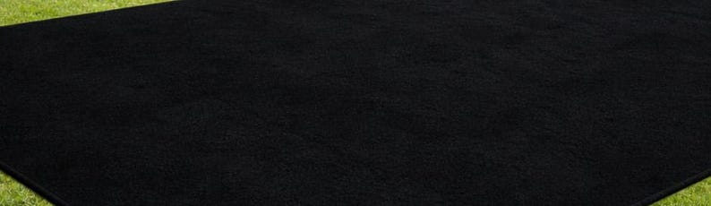 May include: A large, rectangular black rug with a textured surface. The rug is placed on a green surface, creating a contrast in color and texture. The rug's dark color suggests it could be used in various settings.