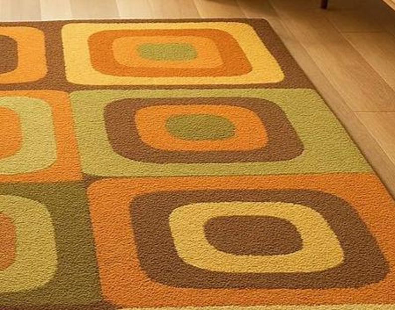 This rug features a retro geometric design with bold, rounded square patterns in warm shades of orange, mustard yellow, olive green, and brown. Its soft texture and mid-century modern style make it a standout piece for any living room or bedroom. The combination of earthy tones and clean, vintage-inspired shapes gives the rug a cozy yet stylish appeal — perfect for adding warmth and personality to your space.