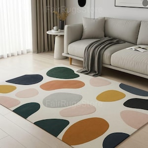 May include: A cream-colored area rug with large, colorful, abstract shapes in shades of green, orange, yellow, pink, gray, and navy blue. The rug is placed on a light wood floor in a living room setting.