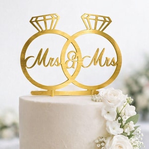 May include: A two-tiered white wedding cake with a gold "Mrs. & Mrs." cake topper. The topper features two interlocking rings with diamond-shaped accents. White roses and greenery cascade down the side of the cake.