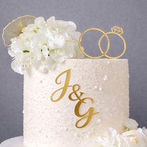 Puede incluir: Una tarta de boda blanca con superficie texturizada, adornada con flores blancas y un adorno de anillos dorados. La tarta presenta las iniciales doradas "J & G". Se encuentra sobre un soporte de madera.
