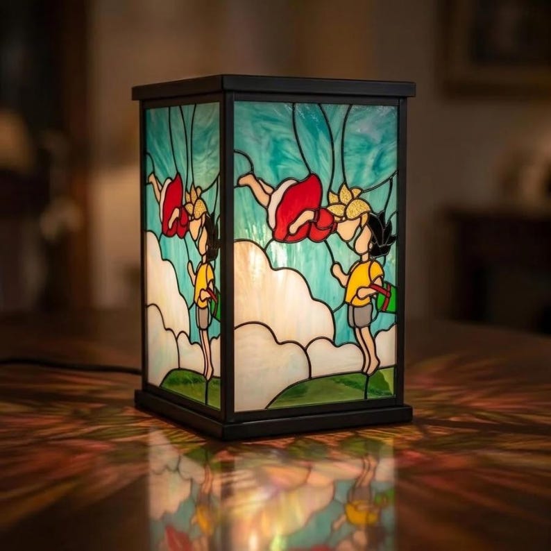 May include: A stained glass lamp with a black metal frame. The lamp features a whimsical design of children floating in the sky with red dresses and yellow shirts. The background is a mix of blue and white, with green at the bottom.