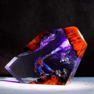 May include: A unique resin sculpture featuring a red, blue, and silver robot figure in a dynamic pose, set against a backdrop of fiery orange and purple hues. The sculpture is encased in a clear, geometric block, showcasing intricate details.