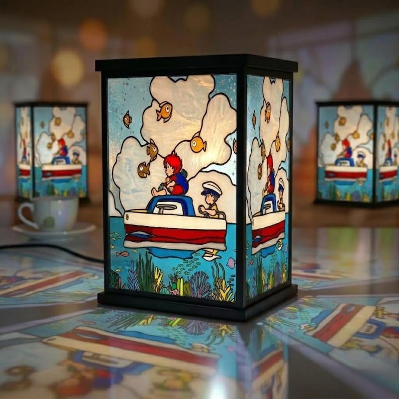 May include: A decorative lamp with a black frame and stained glass panels. The panels depict a cartoon scene of a boat with two people and fish in a blue ocean. The lamp is on a reflective surface, with two other lamps and a teacup in the background.