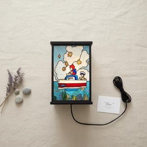 May include: A black framed lamp with a colorful illustration of a boat on the water with two people. The lamp has a red and white boat with a blue sky background and yellow fish. The text "Rain" is on a white card.